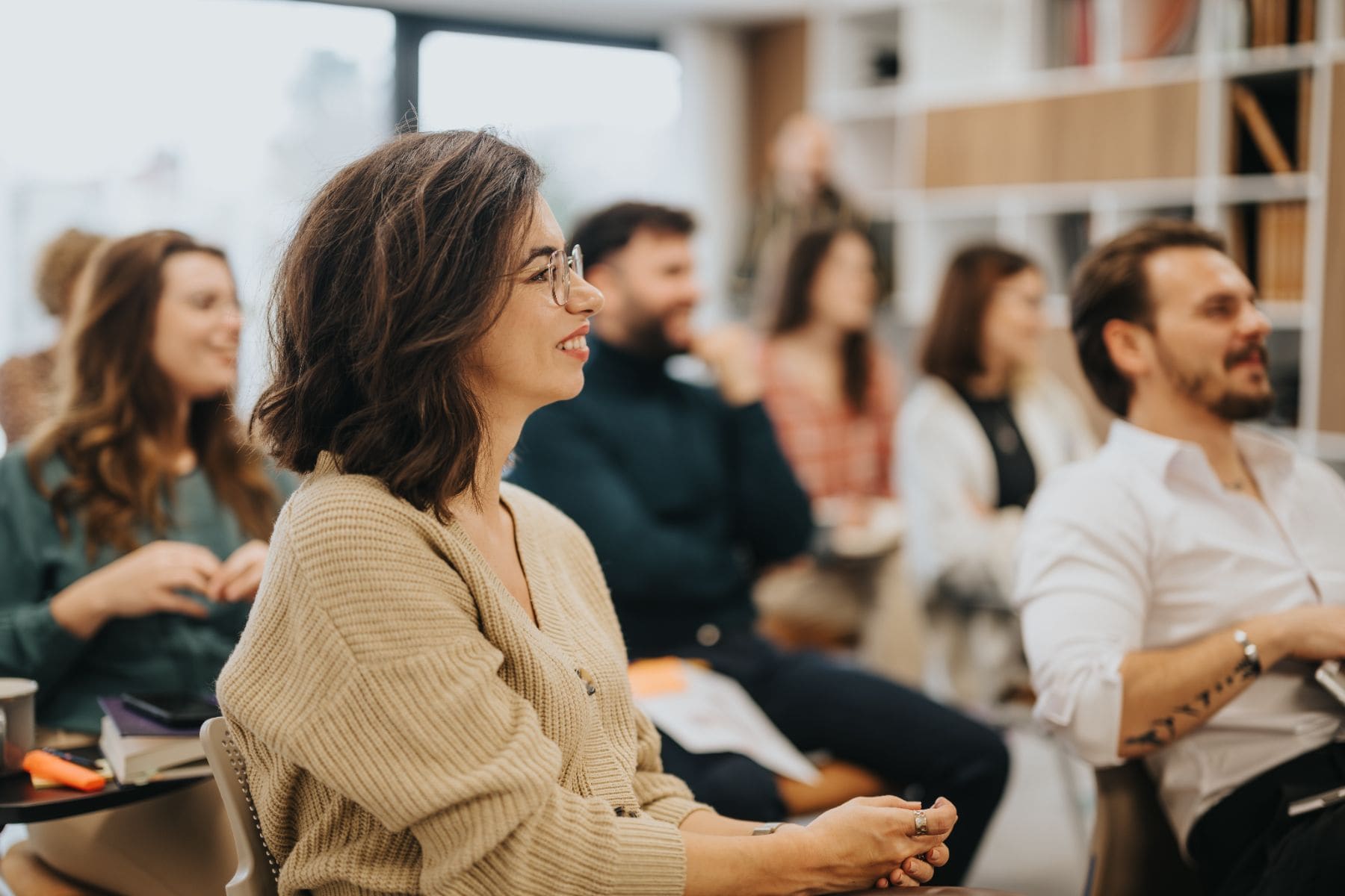 Woman in glasses and beige blazer smiling during indoor business meeting with colleagues