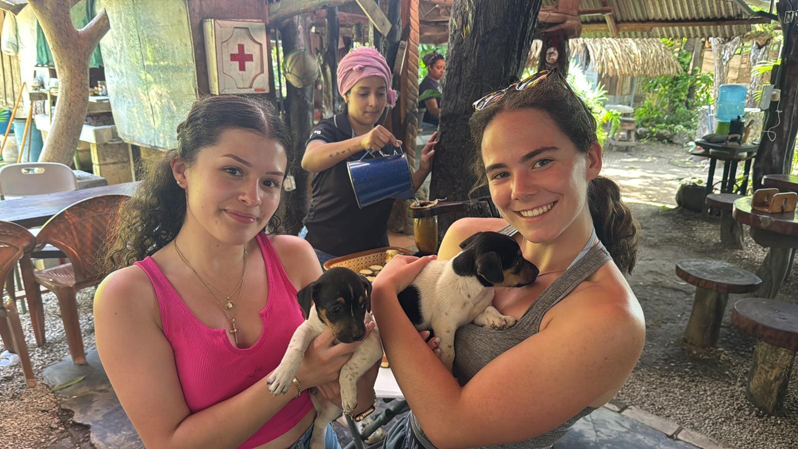 Two young women holding small black and white puppies in an outdoor tropical setting