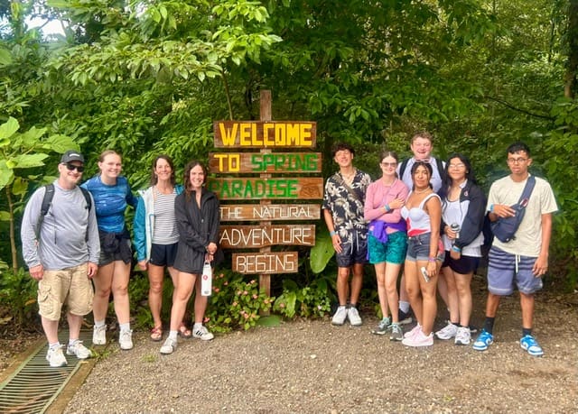 Group of tourists posing together at Spring Paradise nature entrance sign surrounded by green foliage