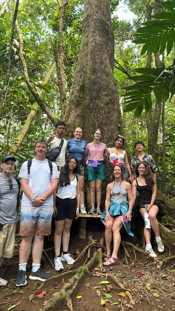 Group of students posing together at base of massive tree trunk in tropical forest