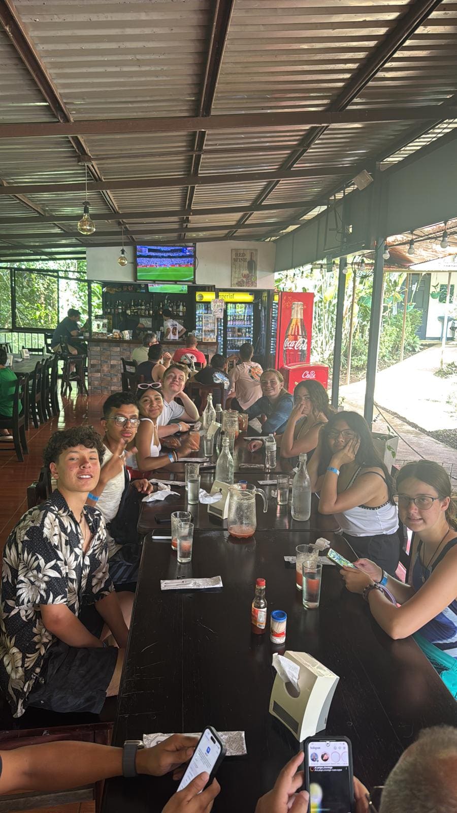 Group of people gathered around a long table at an outdoor covered bar venue watching sports on TV