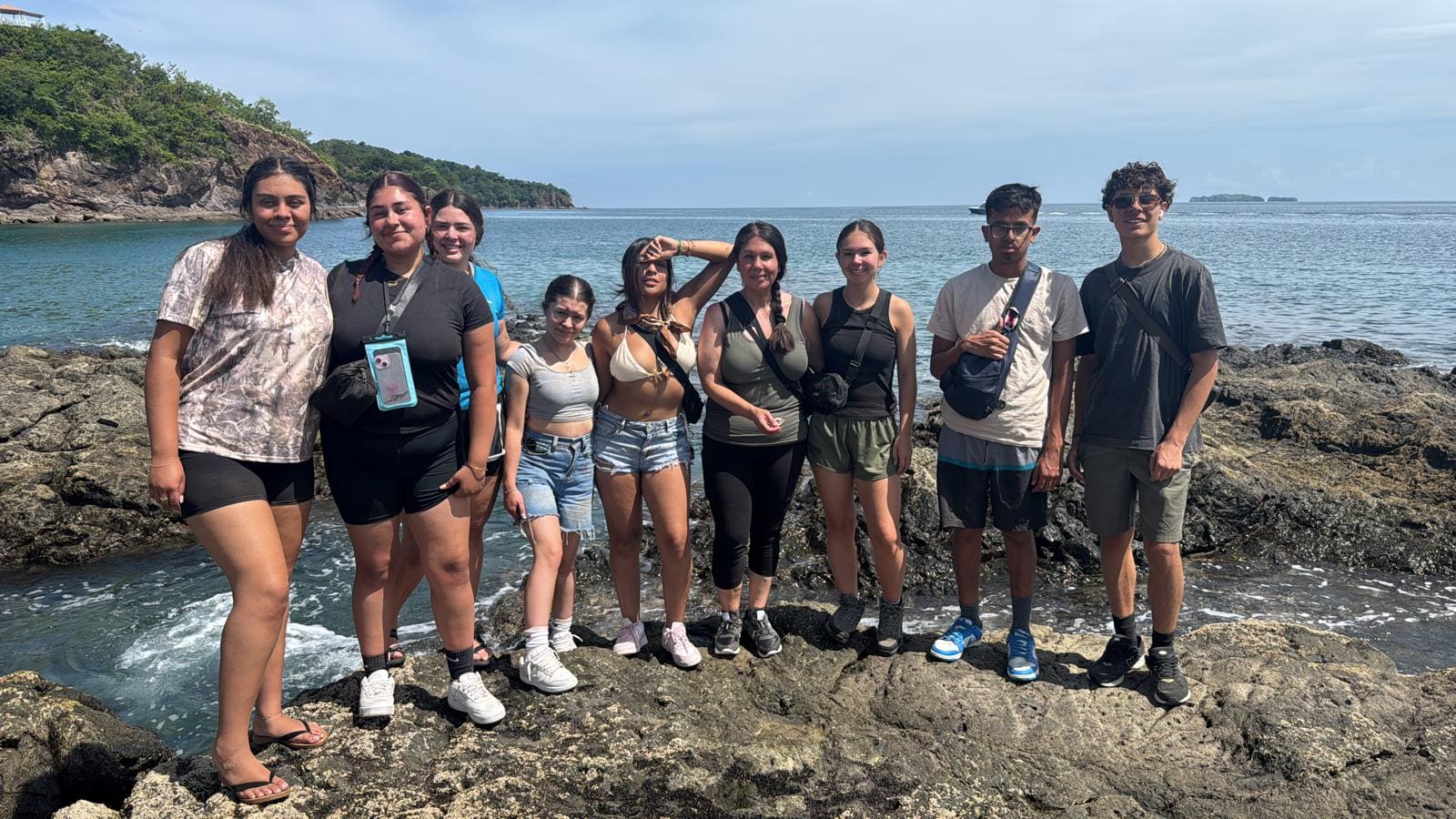 Group of nine young people standing together on rocky beach with ocean and forested coastline behind them
