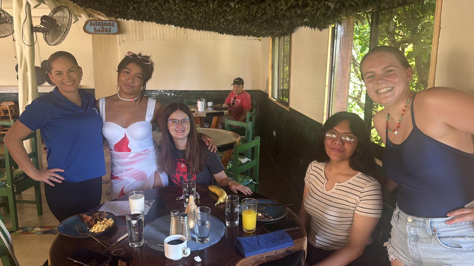 Five women smiling at a dining table in a restaurant with tropical decor and green chairs