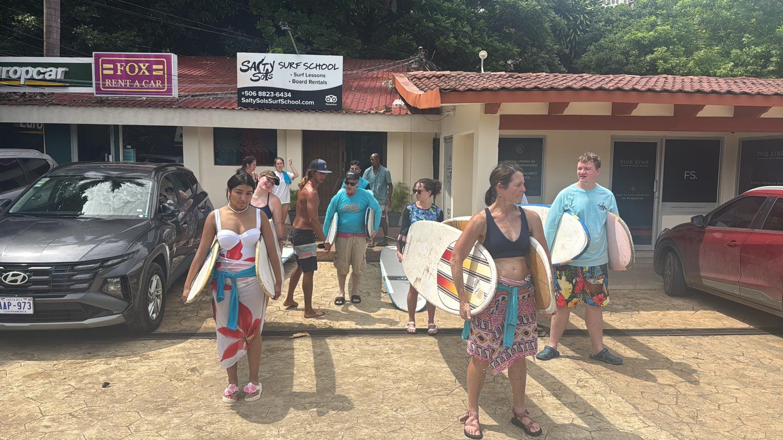 Group of surfers holding boards outside Salty Sols Surf School location with parked cars and storefronts