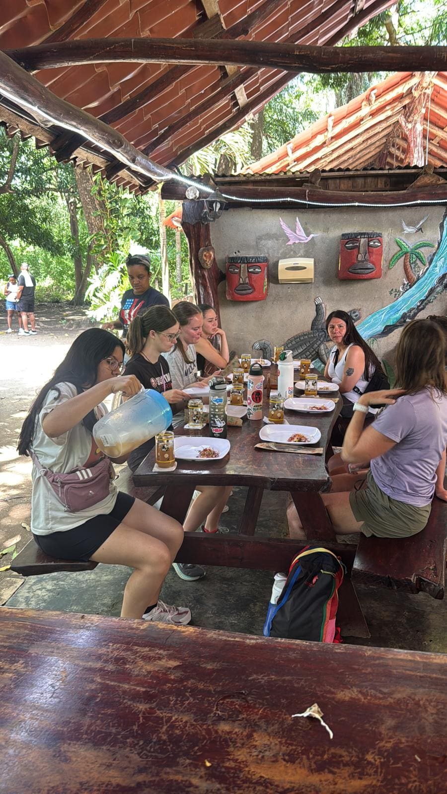 Group of women dining together at rustic outdoor table with indigenous mask decorations on wall