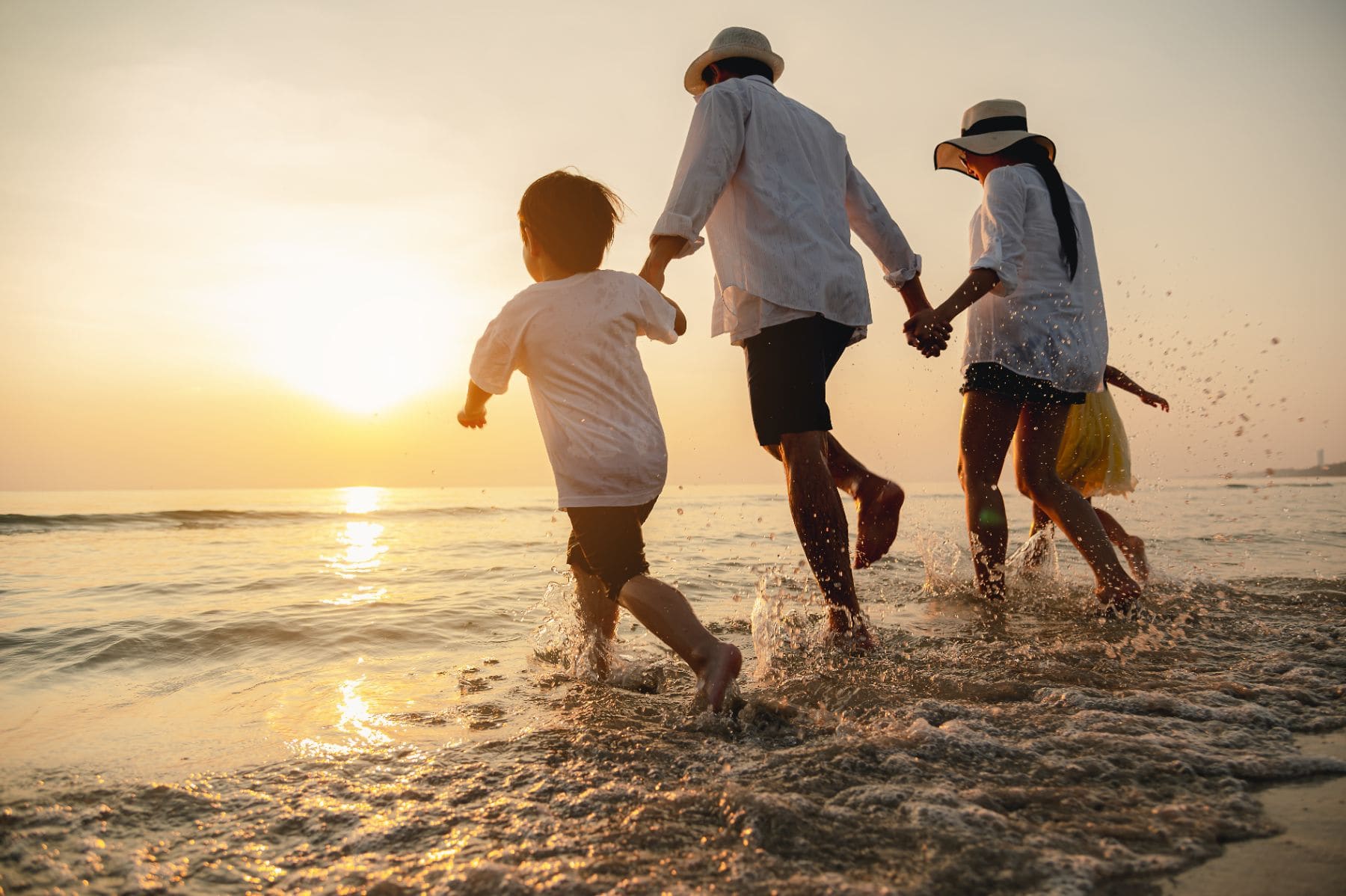 Family of three running into ocean at sunset wearing hats and light clothing