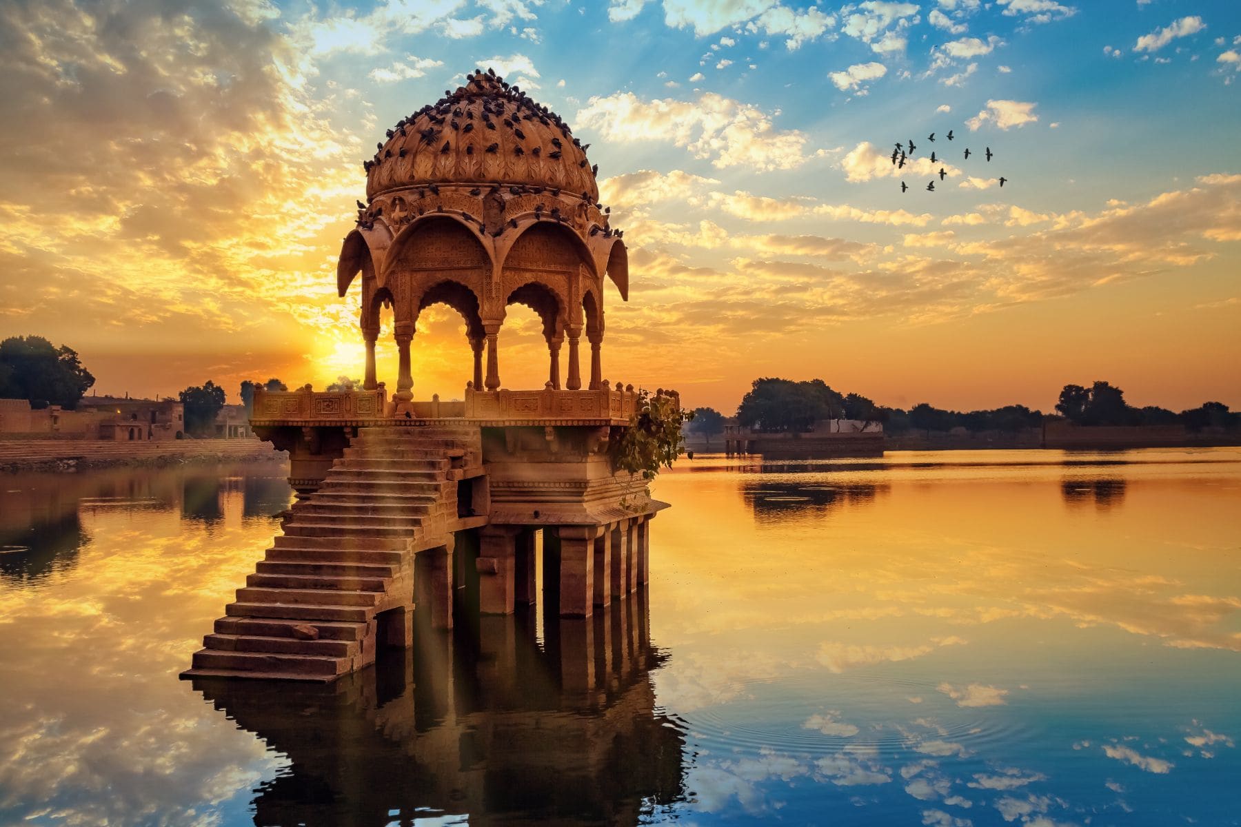 Historic domed pavilion on water at sunset with golden sky and birds flying overhead