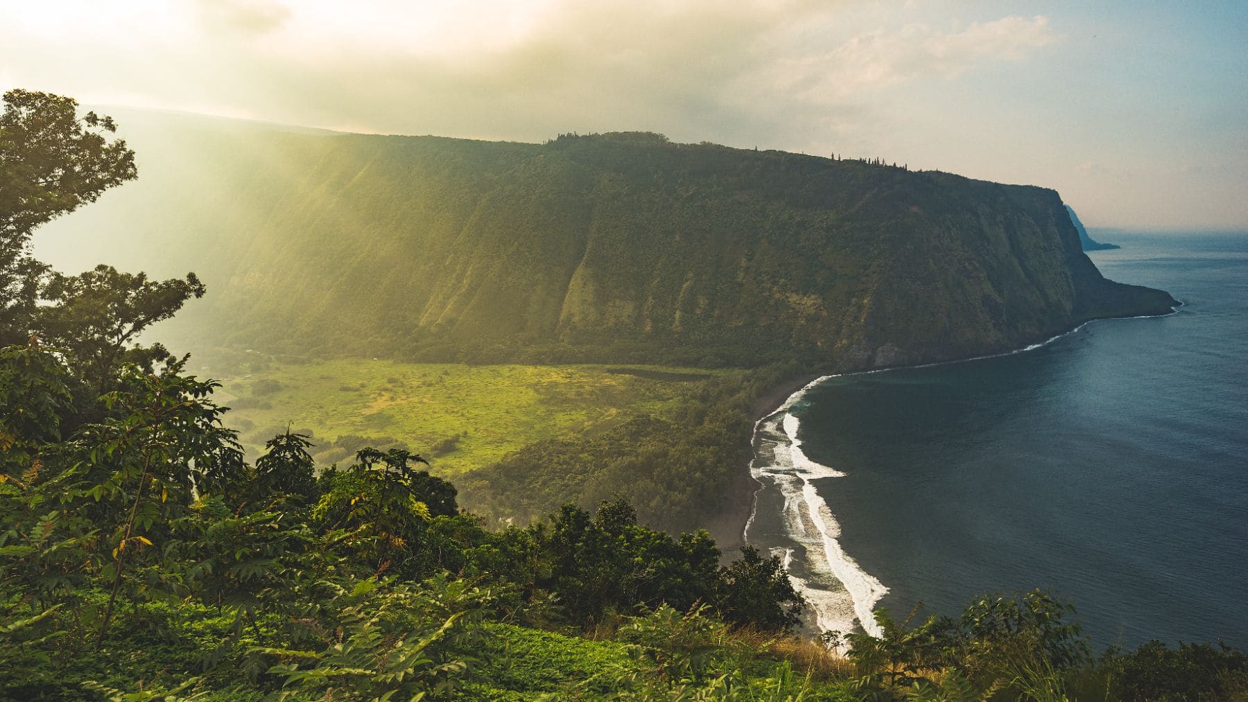 Lush green valley with dramatic sea cliffs and white sand beach along turquoise ocean