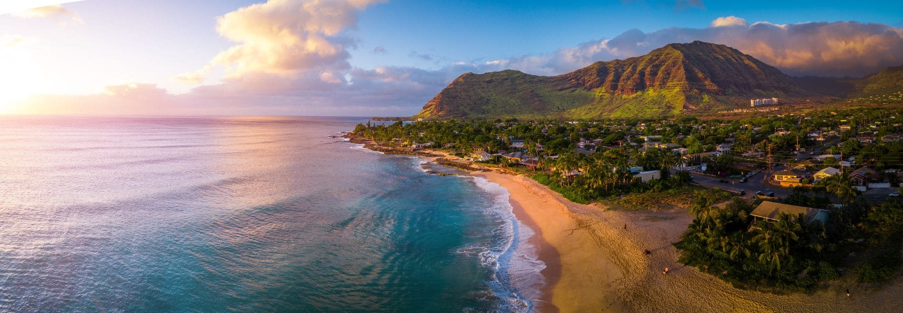 Aerial view of Hawaiian coastal town with golden beach, turquoise waters, palm trees, and green mountain at sunset