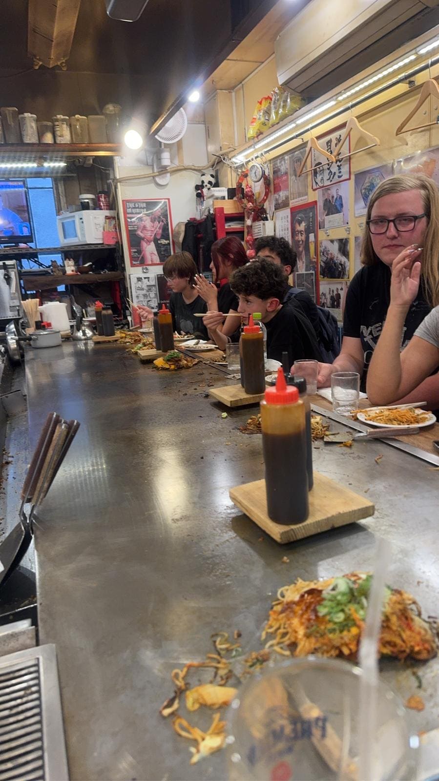 Customers dining at teppanyaki counter with chef cooking noodles and vegetables on griddle