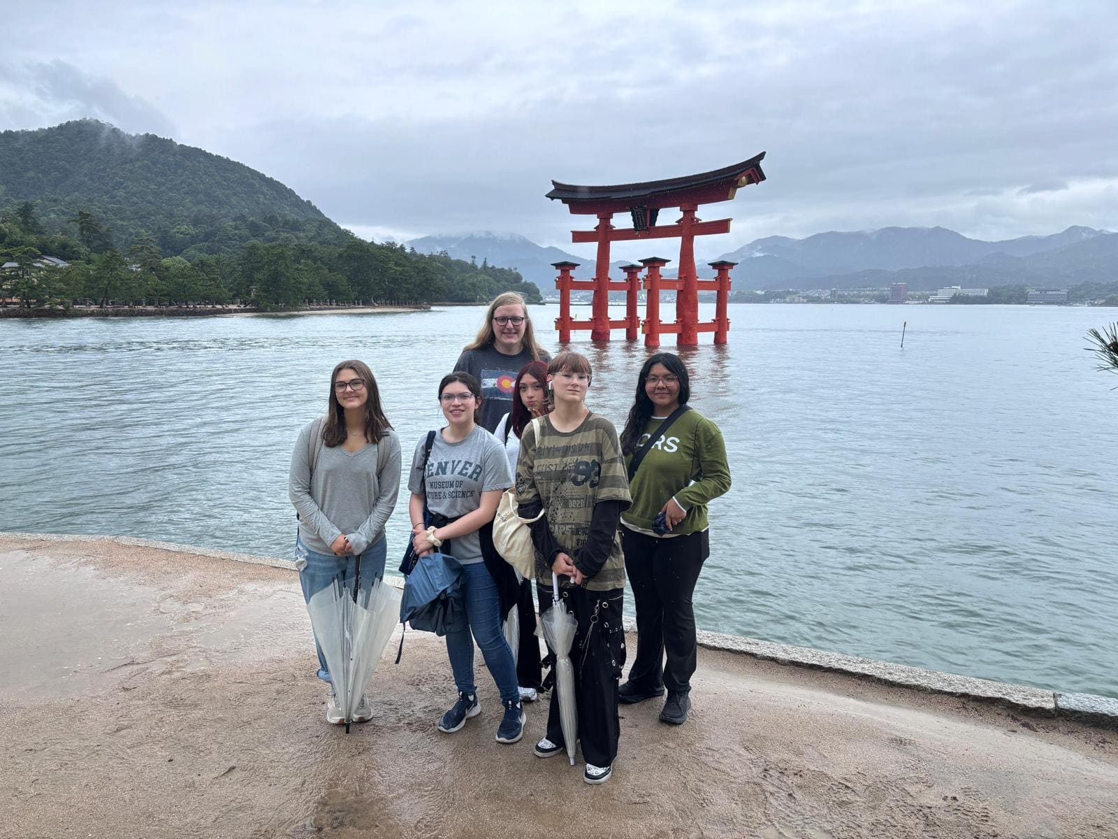 Group of five young people standing on beach with red torii gate in water and mountains behind