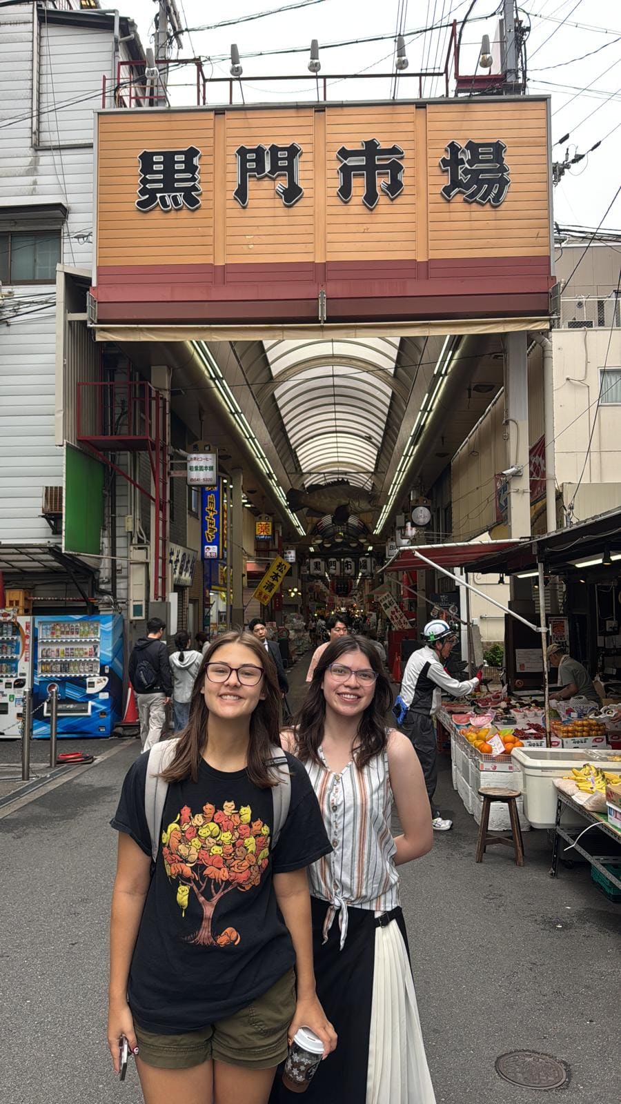 Two young women smiling at entrance of Kuromon Market covered arcade in Osaka, Japan
