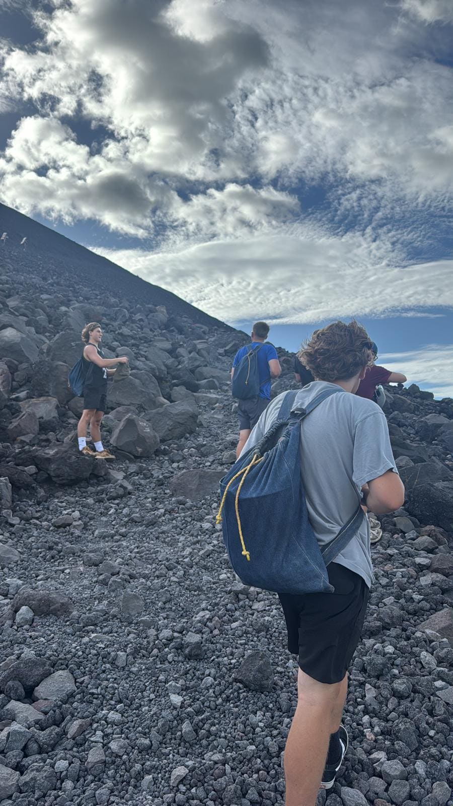 Hikers traversing rocky volcanic terrain with snow-capped mountain and cloudy sky