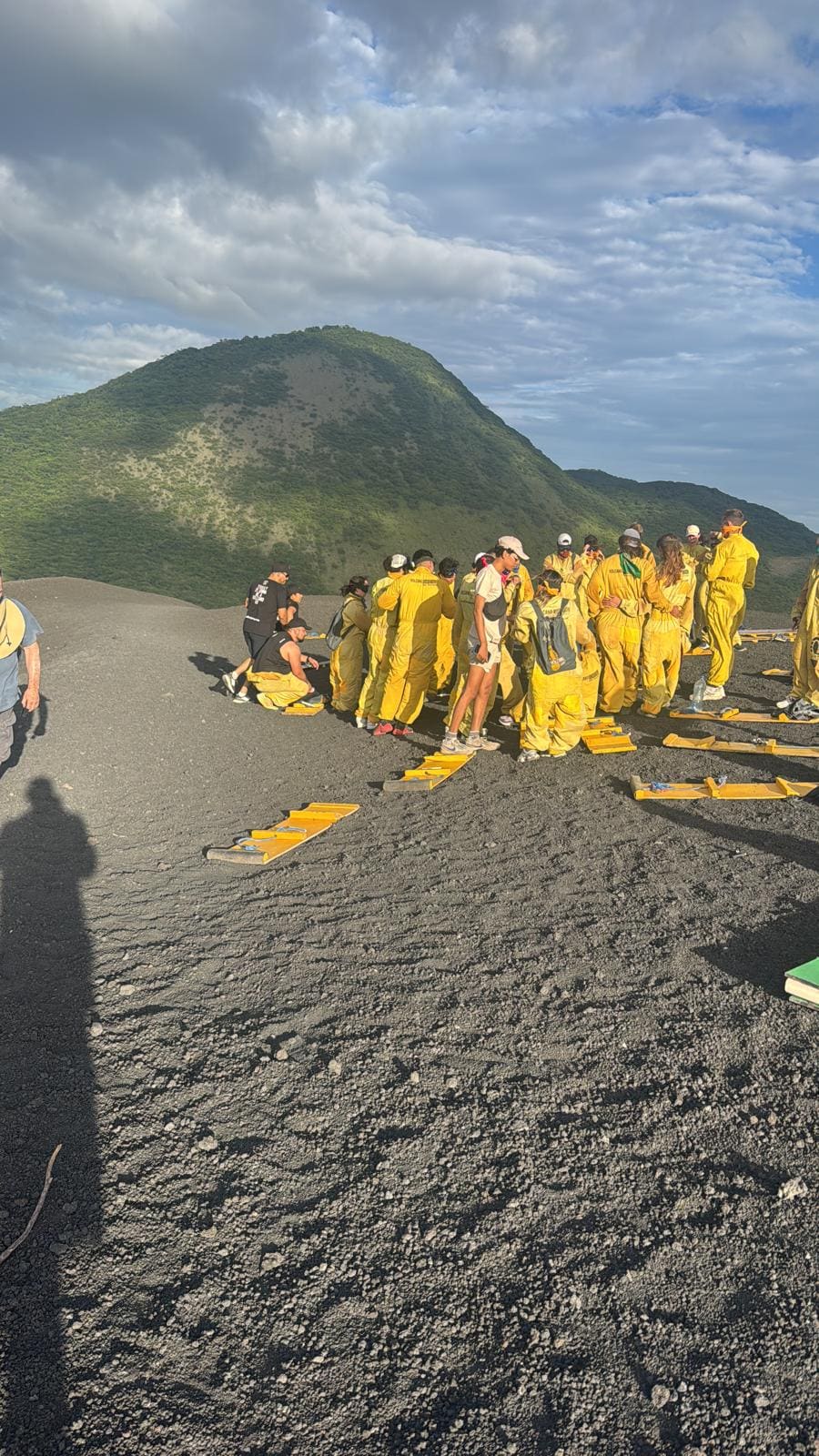Large group wearing yellow safety suits gathered on volcanic ash plain with green cinder cone mountain in background