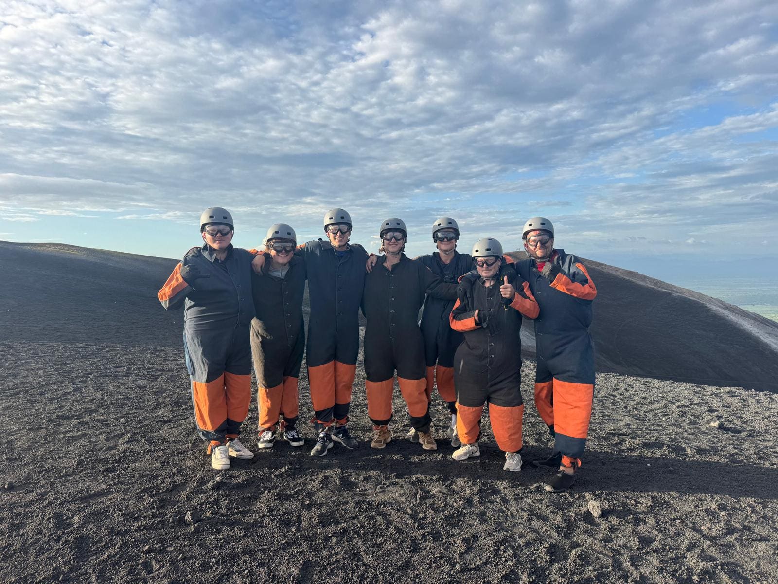 Seven people wearing helmets and black and orange safety gear standing on dark volcanic terrain with mountains in background