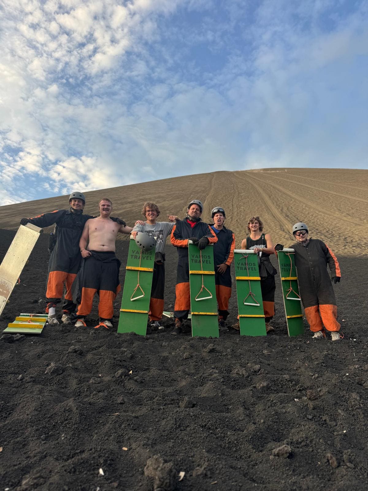 Group of seven people holding green kiteboarding equipment on black sand beach with rolling hills behind them